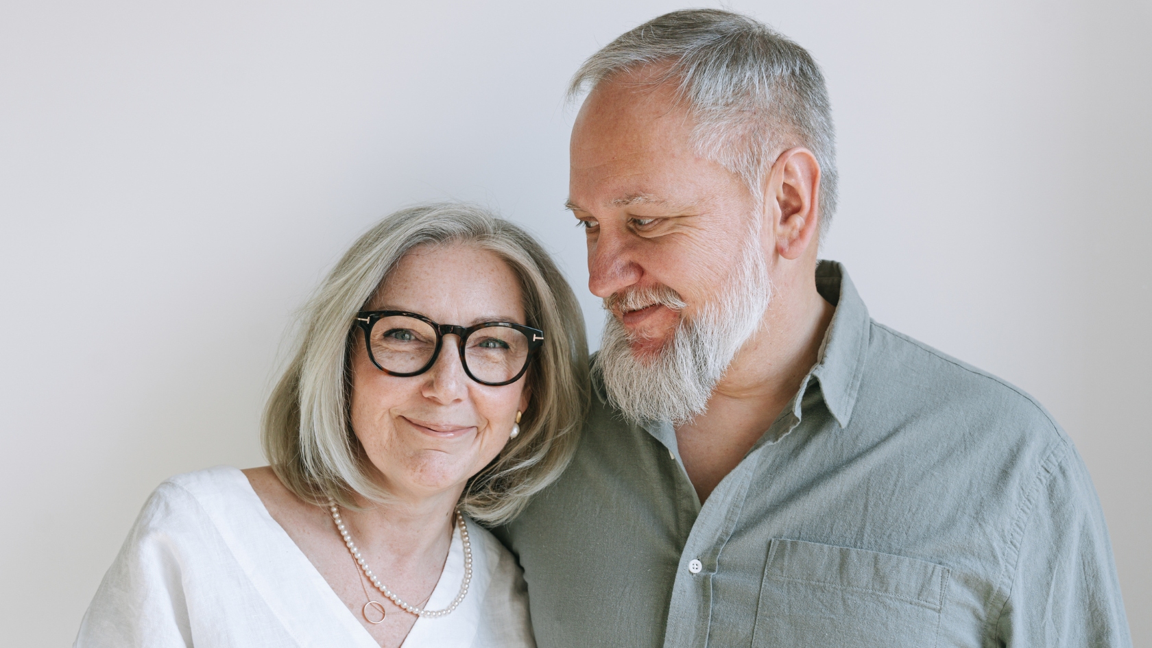 Two older adults standing together, smiling softly against a light background.